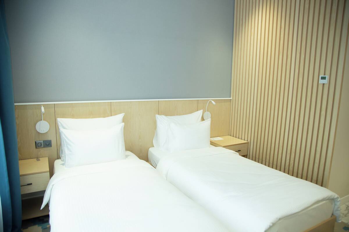 A hotel room with two single beds, wooden headboards, bedside tables, and a neutral-colored wall.