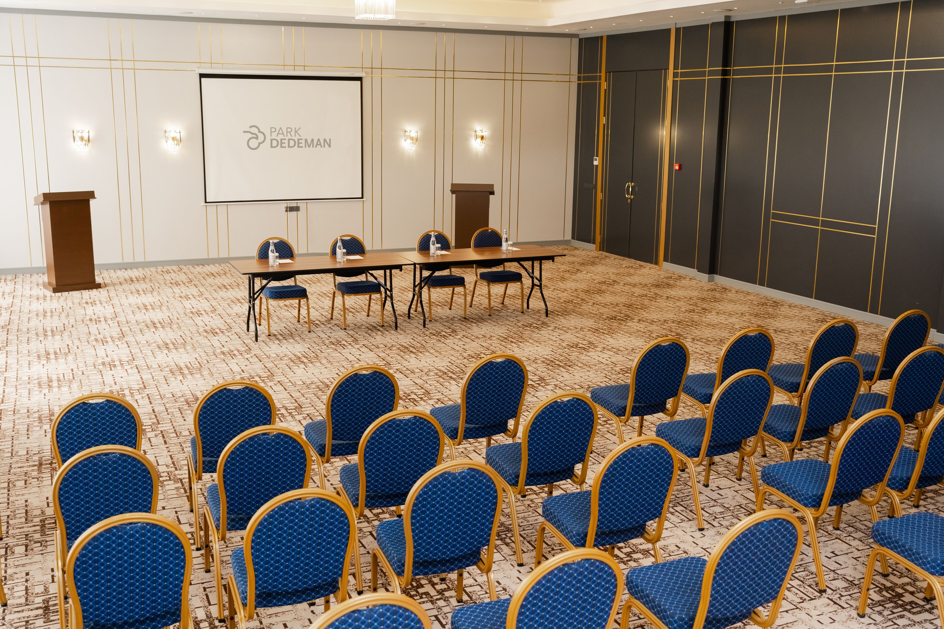 Una sala de conferencias con una larga mesa y sillas azules, una pantalla de proyector y un atril.