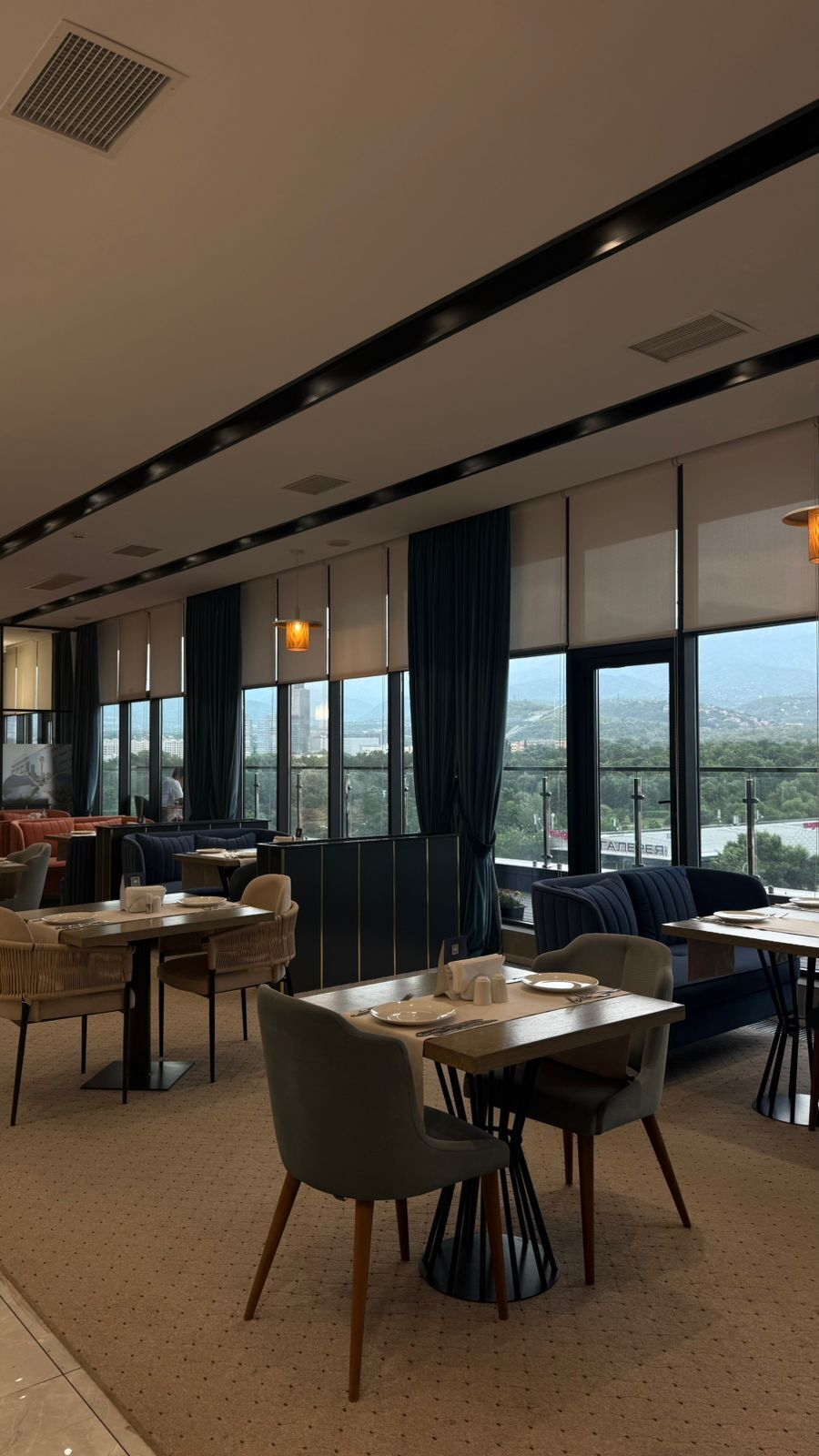 Un comedor moderno con mesas y sillas, ventanas amplias con cortinas y vistas a la ciudad.