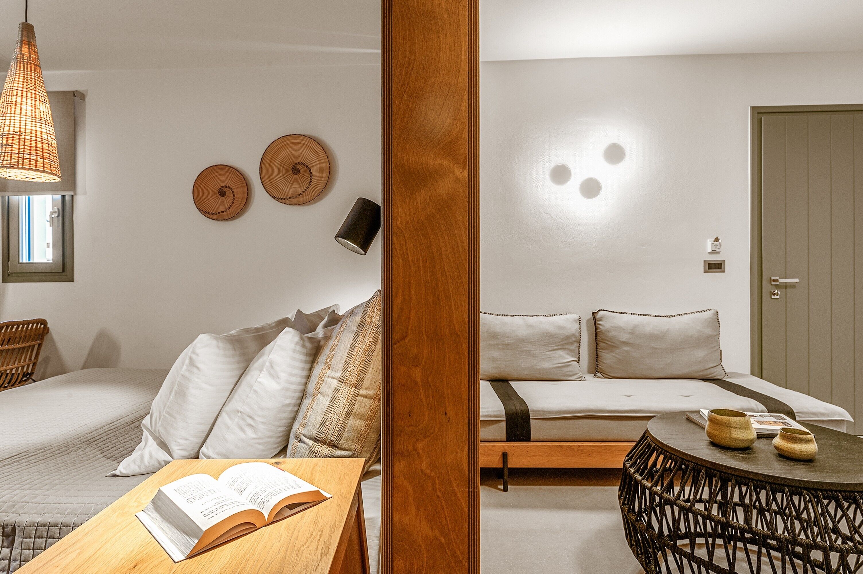 Ein Zimmer mit einem Bett, einem Holztisch mit offenem Buch, einem gewobenen Stuhl und einem runden Tisch mit zwei Vasen.