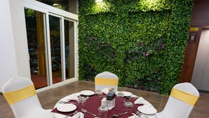 A round dining table with white chairs and a red tablecloth, set against a green leafy wall with a glass door to the left. - Springdale Suites - Nagpur (Nagpur)