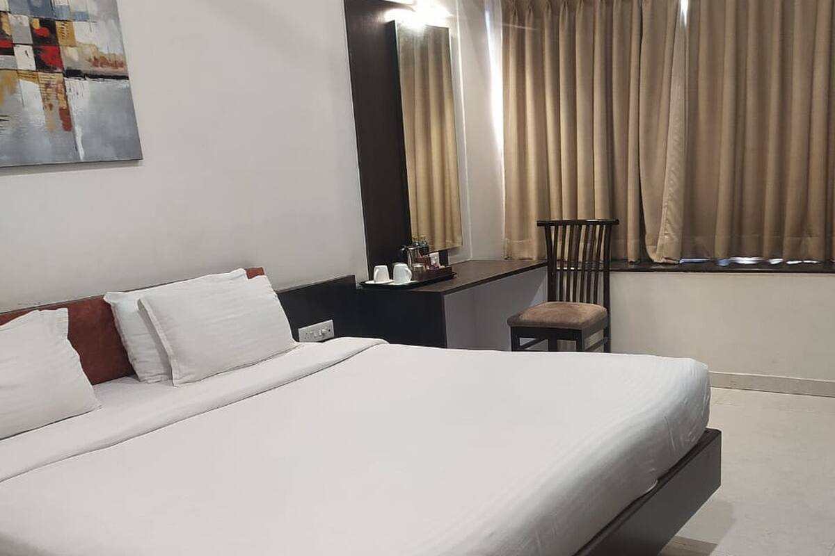 A hotel room with a large bed, a small table with a chair, a window with curtains, and a painting on the wall.