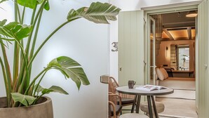 A room with a large potted plant, a round table with chairs, and an open door leading to a dining area.