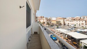 A balcony with a view of a cityscape, a parked car, and a building with a sign. - Studio Apartment "Casa Silvia" with Sea View, Balcony & Air Conditioning (La Maddalena)