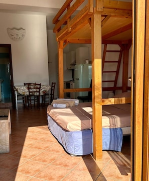 A room with a bunk bed, a dining table with chairs, a kitchen area, and a wooden ladder.