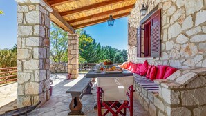 A stone patio with a wooden pergola, a stone wall, a bench with red cushions, and a table with food.