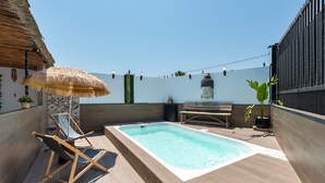 Une piscine avec une terrasse en bois, une chaise longue, un parasol en chaume et un banc.
