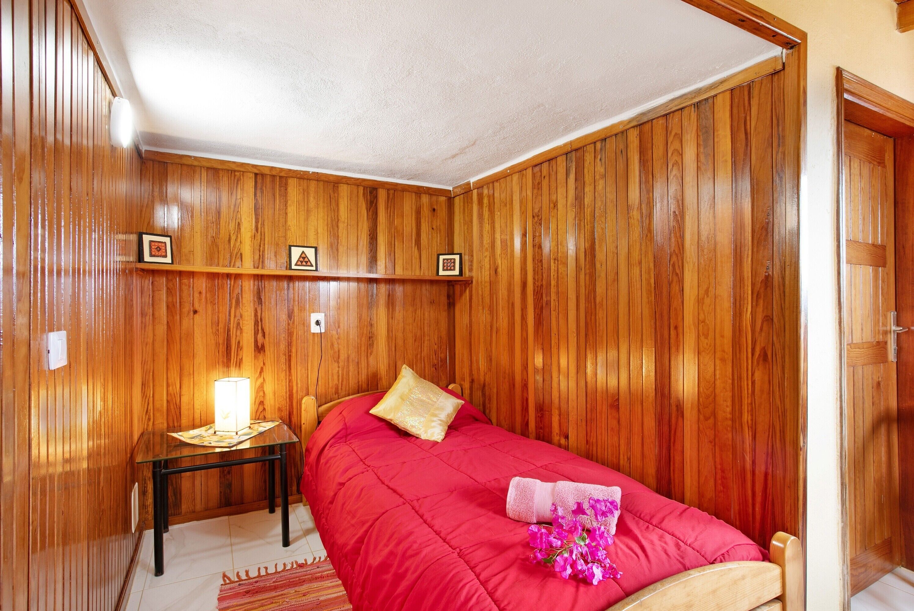 Quarto com painéis de madeira, uma cama, uma mesinha de cabeceira e uma prateleira na parede com quadros.