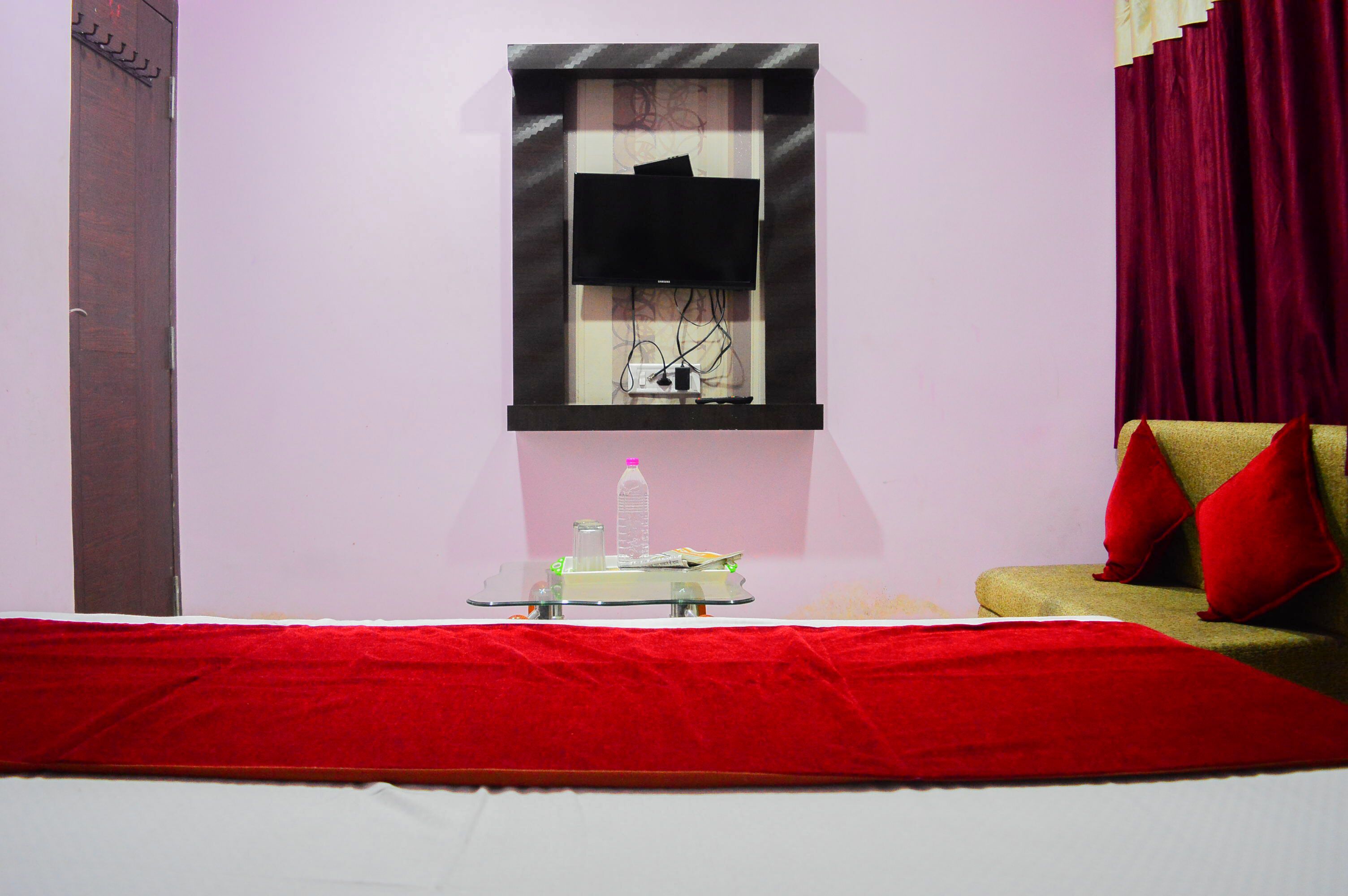 A hotel room with a bed, a small table with a bottle, a TV on the wall, and a red curtain.