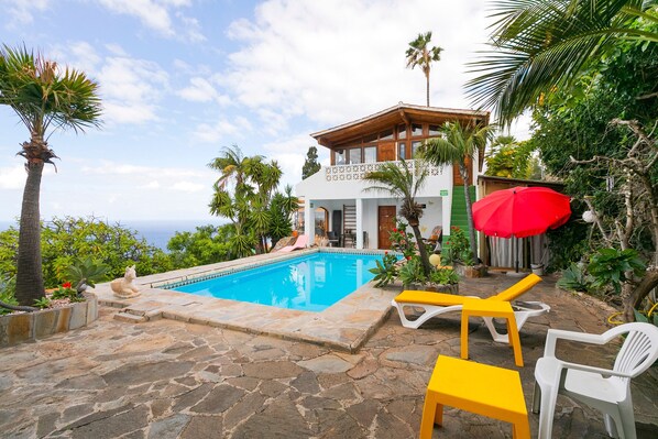 Un espace piscine avec une terrasse en pierre, des chaises longues et un bâtiment doté d’un balcon.
