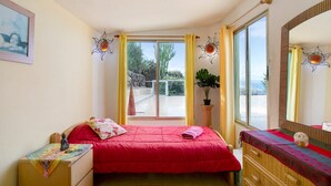 A bedroom with a red bed, a wooden dresser, a large window with curtains, and a potted plant.