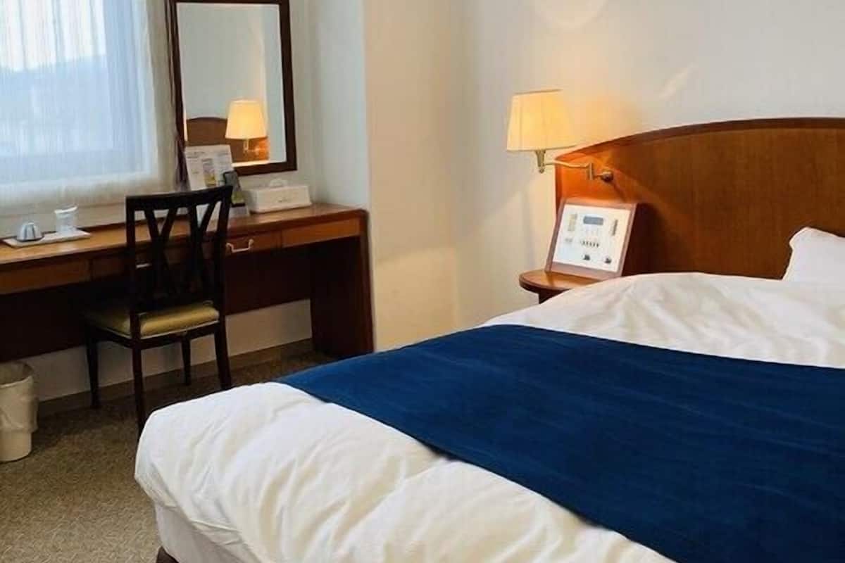 Habitación de hotel con cama, escritorio, silla, lámpara y ventana con cortinas.