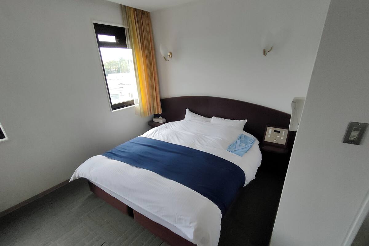 Habitación de hotel con cama, mesita de noche, ventana con cortinas y termostato en la pared.