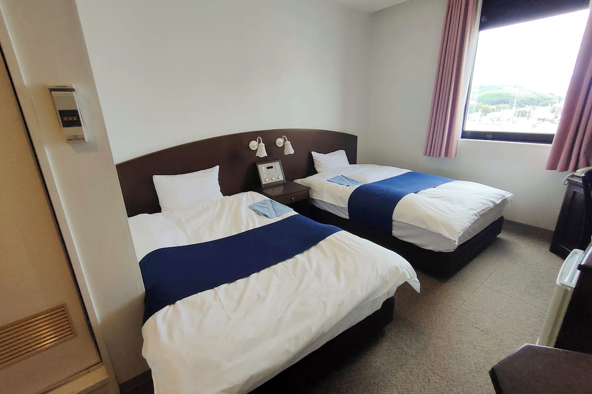 Habitación de hotel con dos camas, una mesita de noche con un reloj, una ventana con cortinas y vistas al exterior.