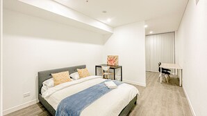 A modern bedroom with a large bed, a small table, and a desk. - Burwood Apartment (Burwood)