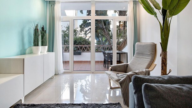 Un salón moderno con sofá, una planta grande y vista a un balcón exterior.