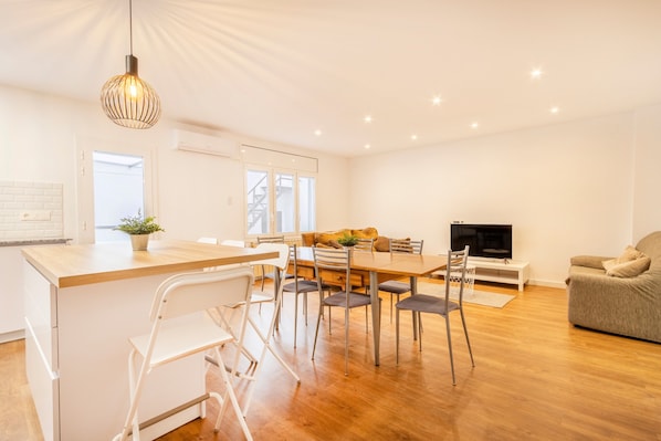 A modern kitchen with a wooden island, white bar stools, a dining table with chairs, a television, and a sofa in a bright living area with wooden flooring.