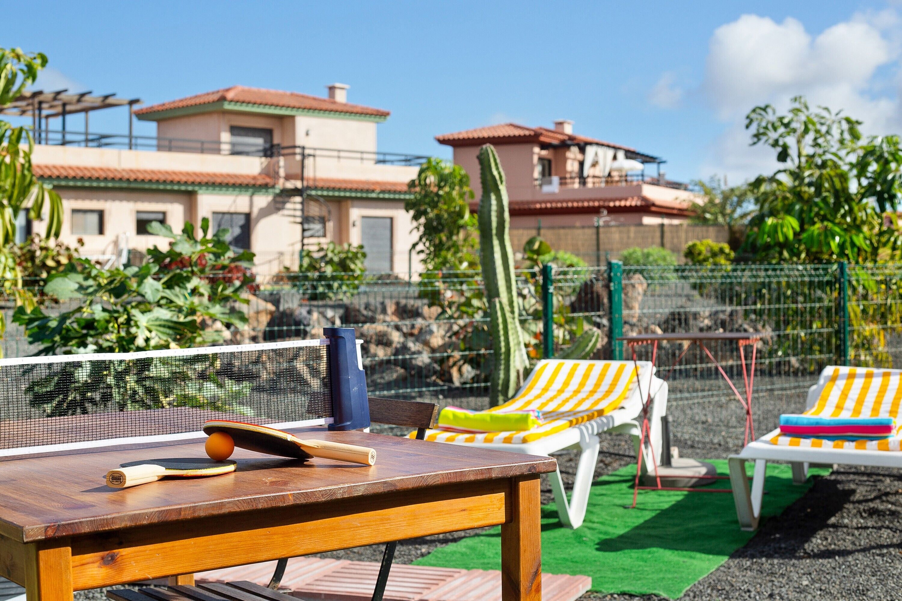 A tennis court with a net, a wooden table with a racket and ball, and two striped lounge chairs.