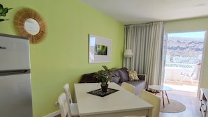 A compact living space with a kitchenette, a dining area with a table and chairs, a sofa, a small round table, and a view of a mountainous landscape through a large window.