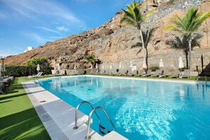 Piscina con un muro de piedra de fondo, sillas de descanso y palmeras.