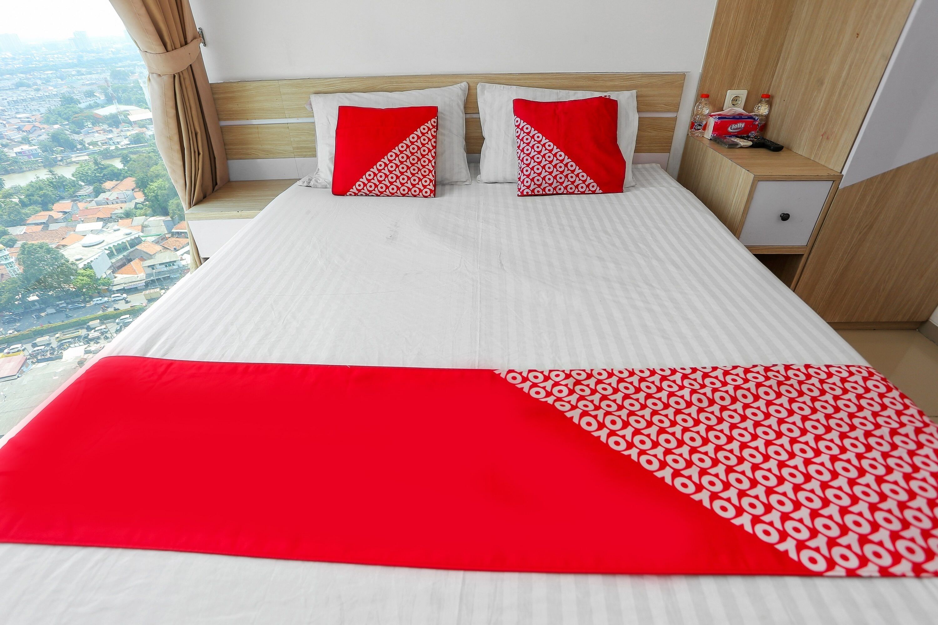 A hotel room with a large bed, two red and white patterned pillows, a red and white throw blanket, a wooden headboard, a nightstand with a bottle and a remote control, and a view of a cityscape through the window.