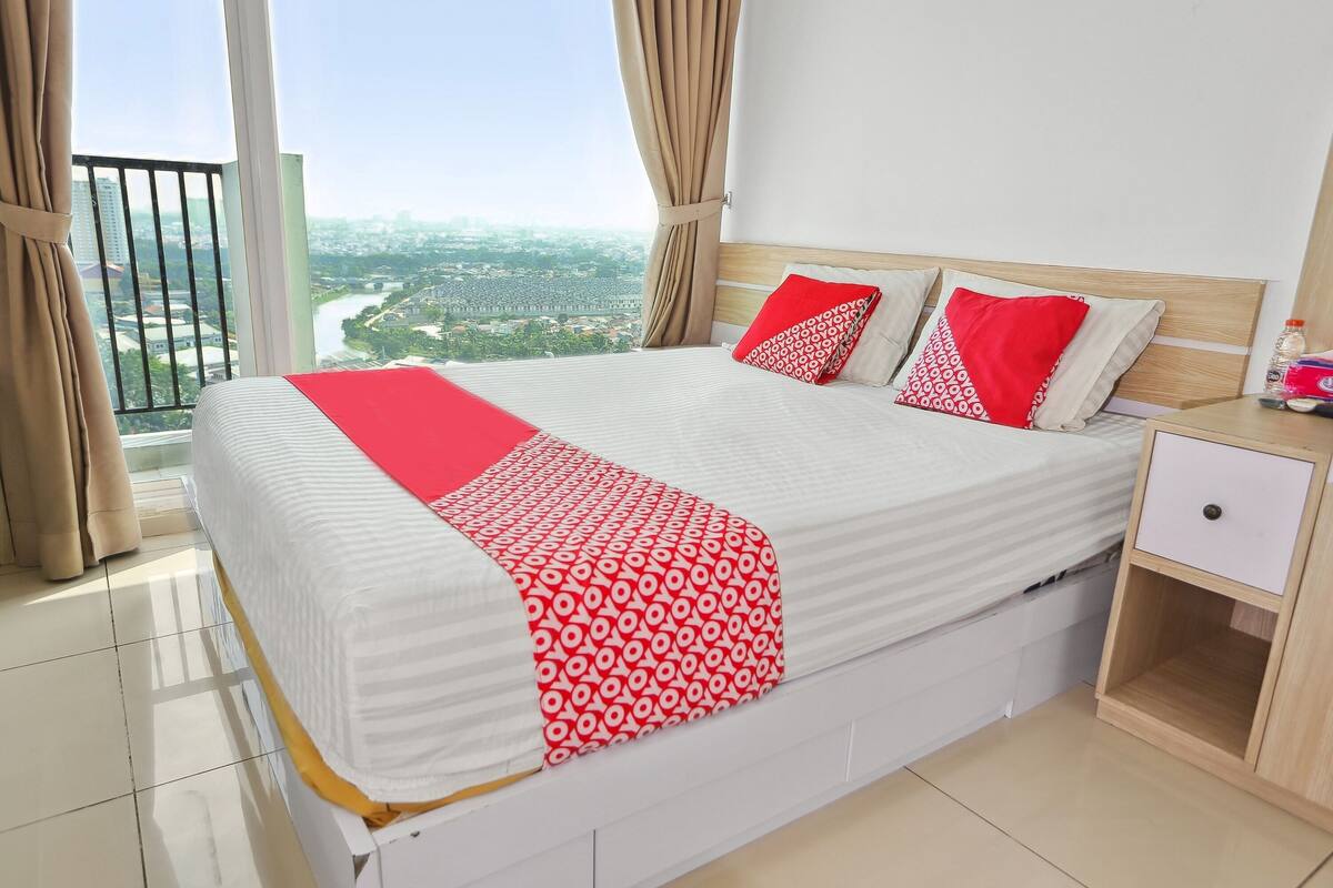 A hotel room with a large bed, a nightstand, and a view of a cityscape through a balcony.