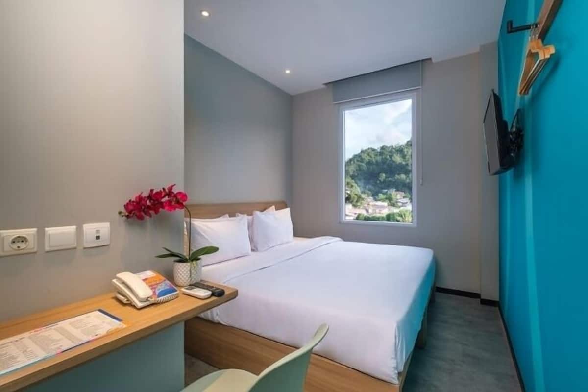 A modern hotel room with a bed, a desk with a phone and papers, a window with a view of greenery, and a blue accent wall.