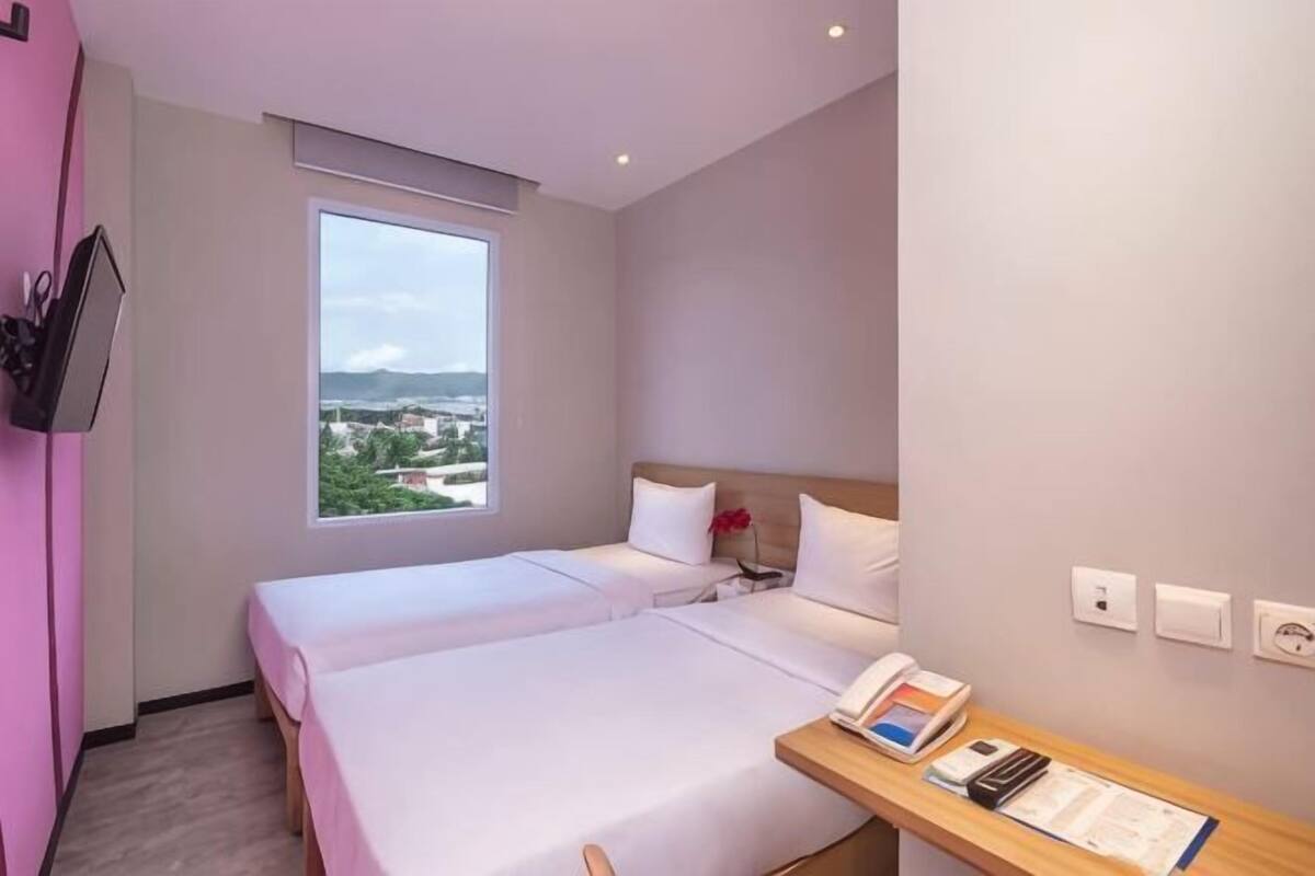 A hotel room with two beds, a desk with a phone and papers, a window with a view, and a TV mounted on the wall.