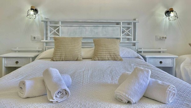 Quarto com cama branca, duas almofadas, cabeceira e pintura pendurada na parede retratando uma praia.