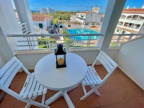 A balcony with a white table and chairs, a lantern, and a view of a swimming pool and buildings.