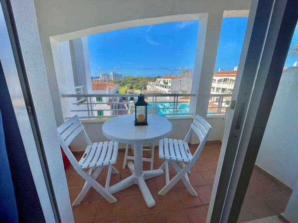 A balcony with a round table and two chairs, overlooking a pool and buildings.