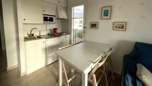 Une cuisine compacte avec des meubles blancs, un four à micro-ondes et une machine à café. On y trouve une table à manger avec des chaises, ainsi qu’un canapé bleu dans l’espace salon.