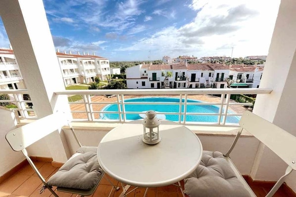 A balcony with a white table and chairs, overlooking a swimming pool and a row of white buildings.