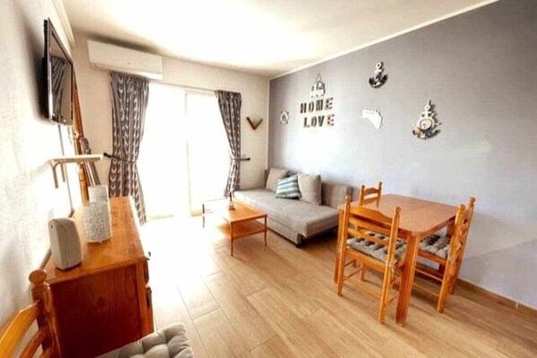 Een woonkamer met een bank, een houten eettafel met stoelen, een klein houten bijzettafeltje en een televisie op een standaard.