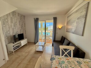A modern living room with a flat-screen TV, a sofa, a dining table, and a balcony with a pool view.