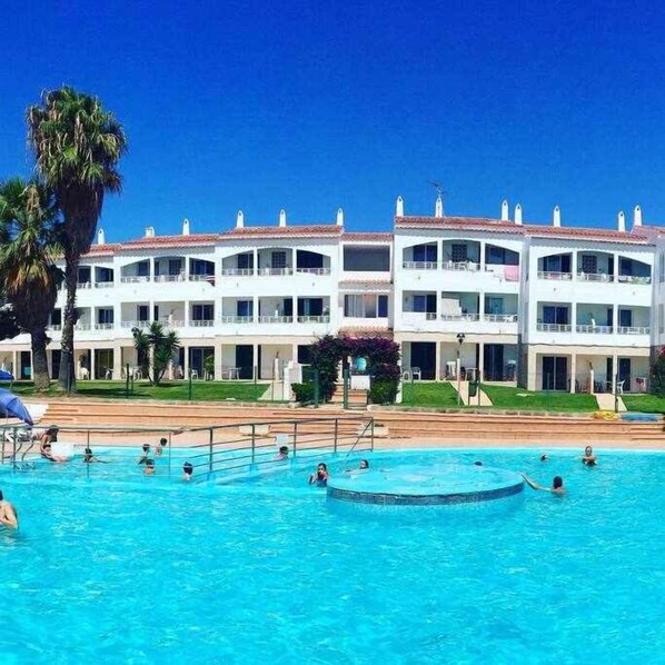 A hotel with a large swimming pool, multiple balconies, and a clear blue sky.