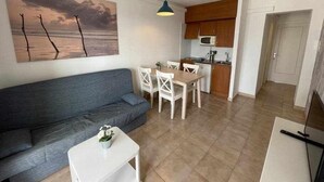 A living room with a gray sofa, a white coffee table, and a painting of a sunset. There is a kitchen area with wooden cabinets and a microwave.