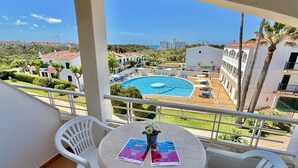 A balcony with a view of a swimming pool, white chairs, and a table with brochures.