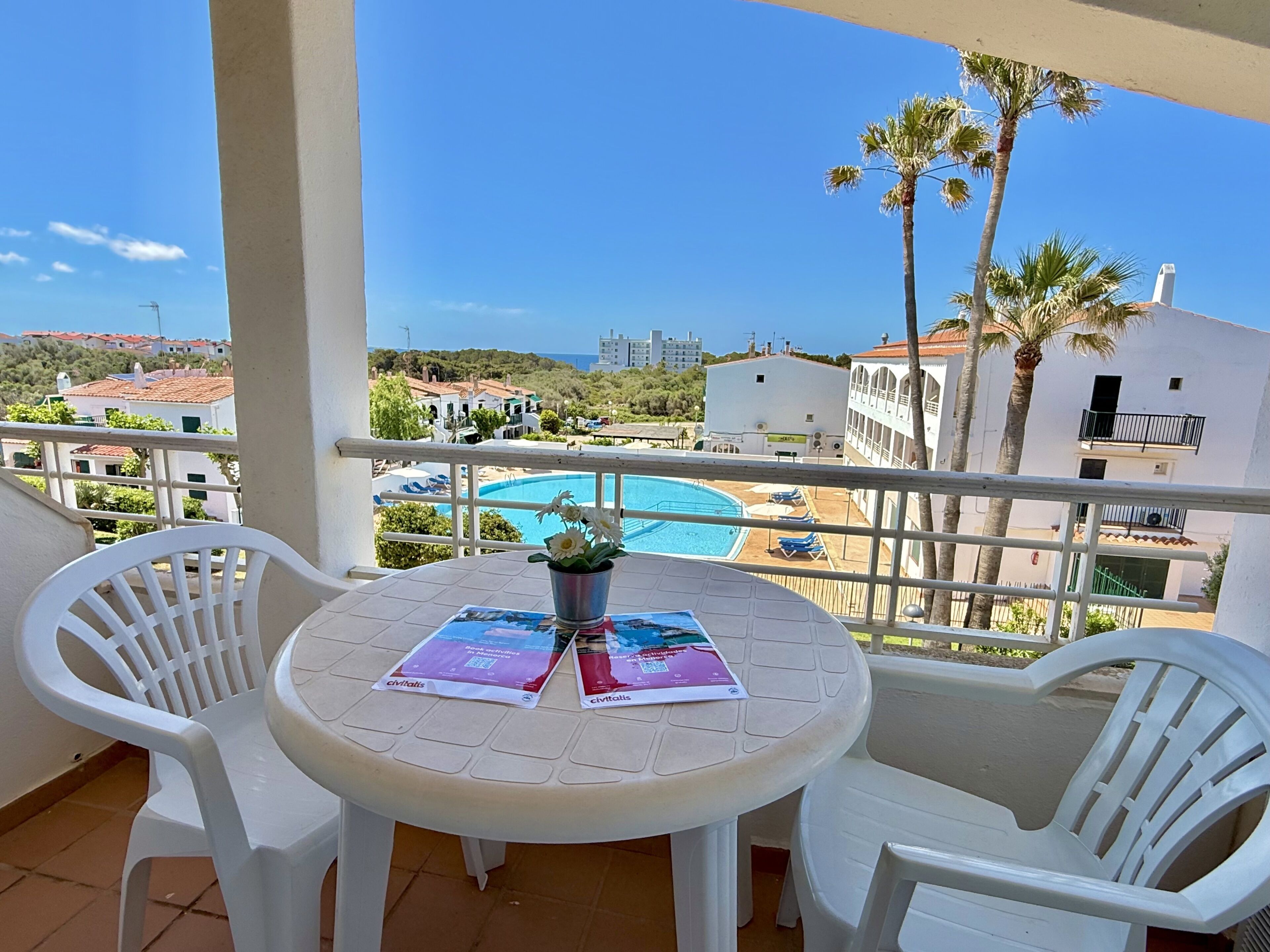 A balcony with a view of a swimming pool, white chairs, and a table with brochures.