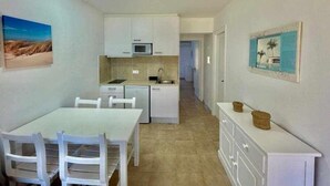 Una cocina compacta con armarios blancos, microondas y lavamanos. Cuenta con una mesa de comedor con sillas y un mueble bajo con cestas.