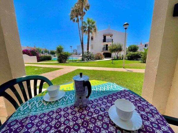Une table dressée pour deux personnes, recouverte d’une nappe à motifs, avec une théière et deux tasses, donnant sur un jardin bien entretenu et un bâtiment visible au loin.