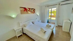 Una habitación de hotel con cama doble, mesita de noche, armario pequeño y ventana con cortinas.