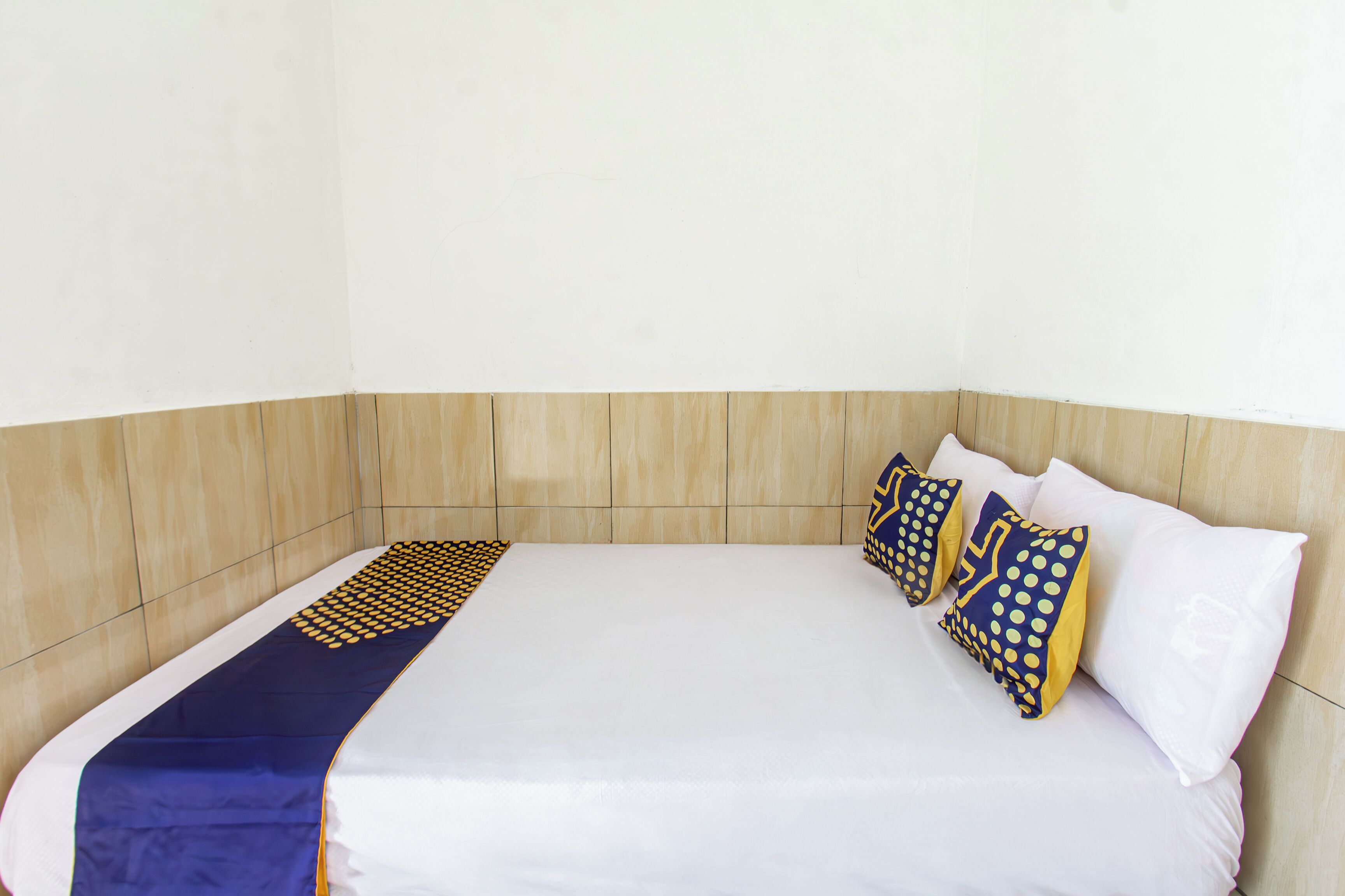 Una cama individual con colchón blanco y manta de diseño azul y amarillo. Dos almohadas con un estampado de lunares azules y amarillos. La habitación tiene un cabecero de madera y una pared blanca lisa.