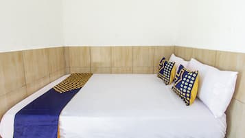 Una cama individual con colchón blanco y manta de diseño azul y amarillo. Dos almohadas con un estampado de lunares azules y amarillos. La habitación tiene un cabecero de madera y una pared blanca lisa.