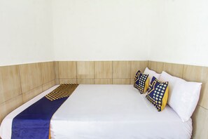 Un llit individual amb matalàs blanc i manta de disseny blau i groc. Hi ha dos almoinos amb dibuix de punts blaus i grocs. La habitació té un capçal de llit de fusta i una paret blanca sense adorns.