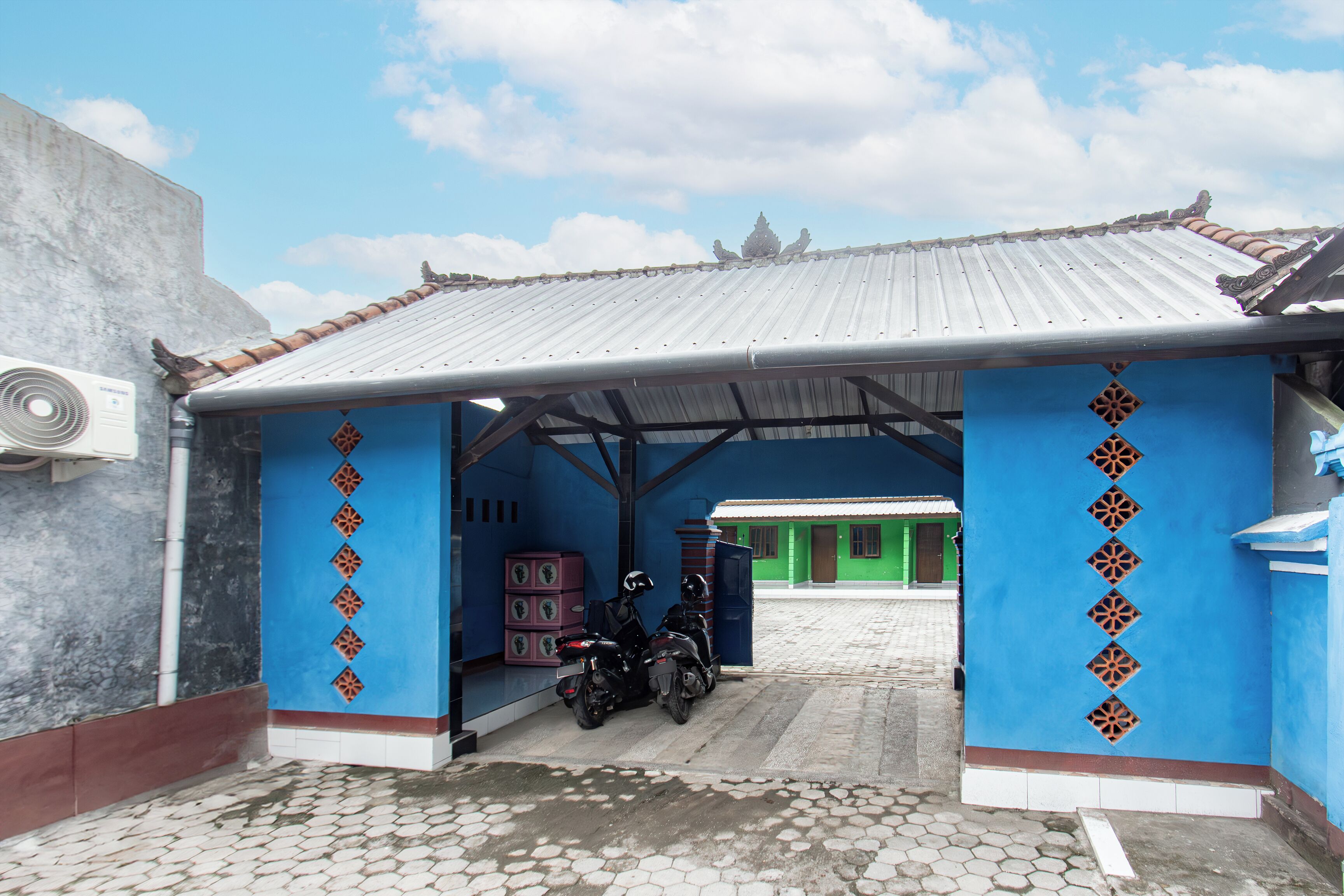 Un espace extérieur abrité où est garée une moto, un bâtiment vert en arrière-plan et une structure bleue ornée de motifs décoratifs.