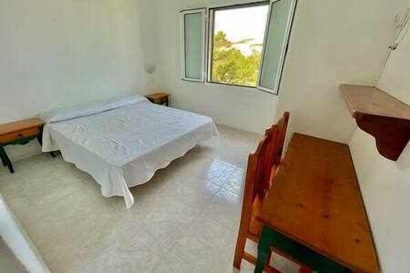 A small, sparsely furnished room with a single bed, a wooden table, and a window with a view of greenery.