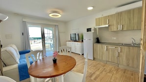 A modern kitchen with wooden cabinets, a white refrigerator, and a built-in microwave. There is a dining area with a wooden table and white chairs. - Gregal - Castell Sol - Arenal de Castell (S'Arenal D'En Castell (Menorca) (Urbanitzacio))