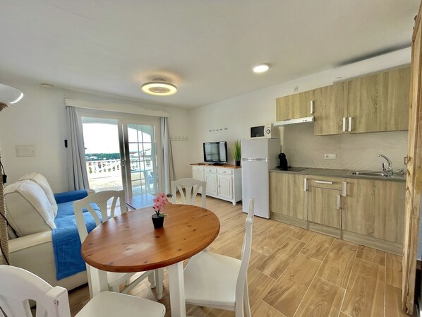 Una cocina moderna con armarios de madera, un refrigerador blanco y un microondas integrado. Hay una zona de comedor con una mesa de madera y sillas blancas.