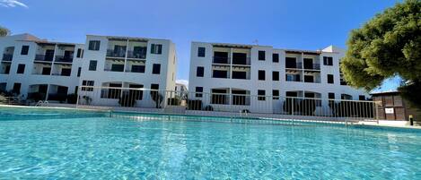 A clear pool with a view of a white building with balconies and a tree to the right.